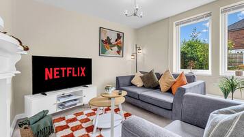 Uma sala de estar com uma televisão exibindo 'NETFLIX', um sofá, uma mesa de centro e vista para o verde lá fora.