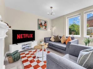 Un salon avec un téléviseur affichant le logo « NETFLIX », un canapé, une table basse et une vue sur la verdure à l’extérieur.