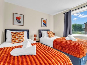 Une chambre d’hôtel avec deux lits, chacun recouvert d’une couverture orange et doté d’oreillers blancs, une table de chevet avec une lampe et une fenêtre donnant sur un paysage verdoyant.