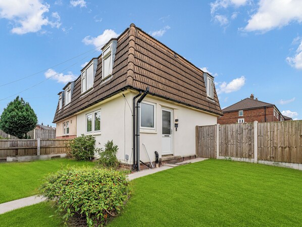 Une maison à deux étages avec un toit en pente, une porte d’entrée et un petit jardin devant, bordé d’une haie.