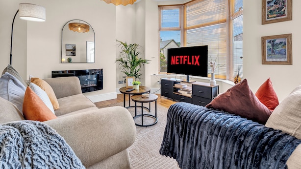 A modern living room with a television displaying 'NETFLIX,' a sofa, a coffee table, and a large window with blinds. - A terraced cottage on Leek Road, positioned for Buxton and the Peak District. (Buxton)
