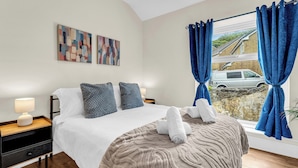 A hotel room with a bed, two pillows, a bedside table with a lamp, a window with curtains, and artwork on the wall. - A terraced cottage on Leek Road, positioned for Buxton and the Peak District. (Buxton)