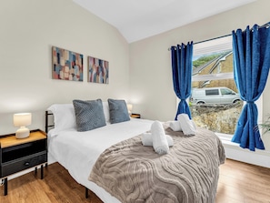Habitación de hotel con una cama, dos almohadas, una mesita de noche con lámpara, una ventana con cortinas y una obra de arte en la pared.
