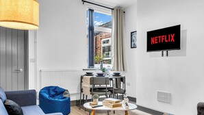 Moderni olohuone, jossa on ruokailutila, suuri ikkuna verhoilla, seinälle asennettu televisio, jossa näkyy teksti ”NETFLIX”, ja viihtyisä istumaryhmä.