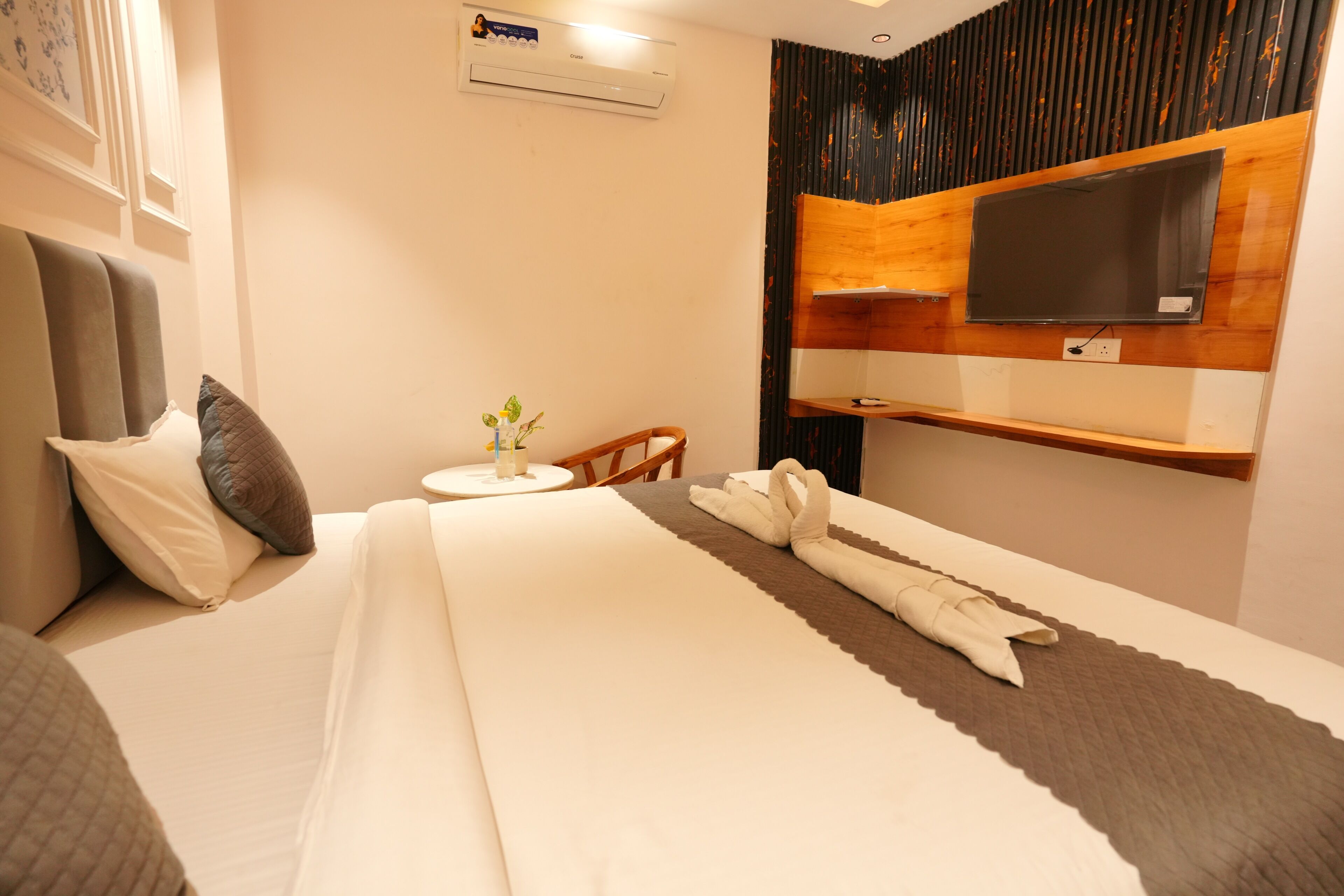 A hotel room with a bed, a small table with a plant, a TV mounted on the wall, and a wooden headboard.