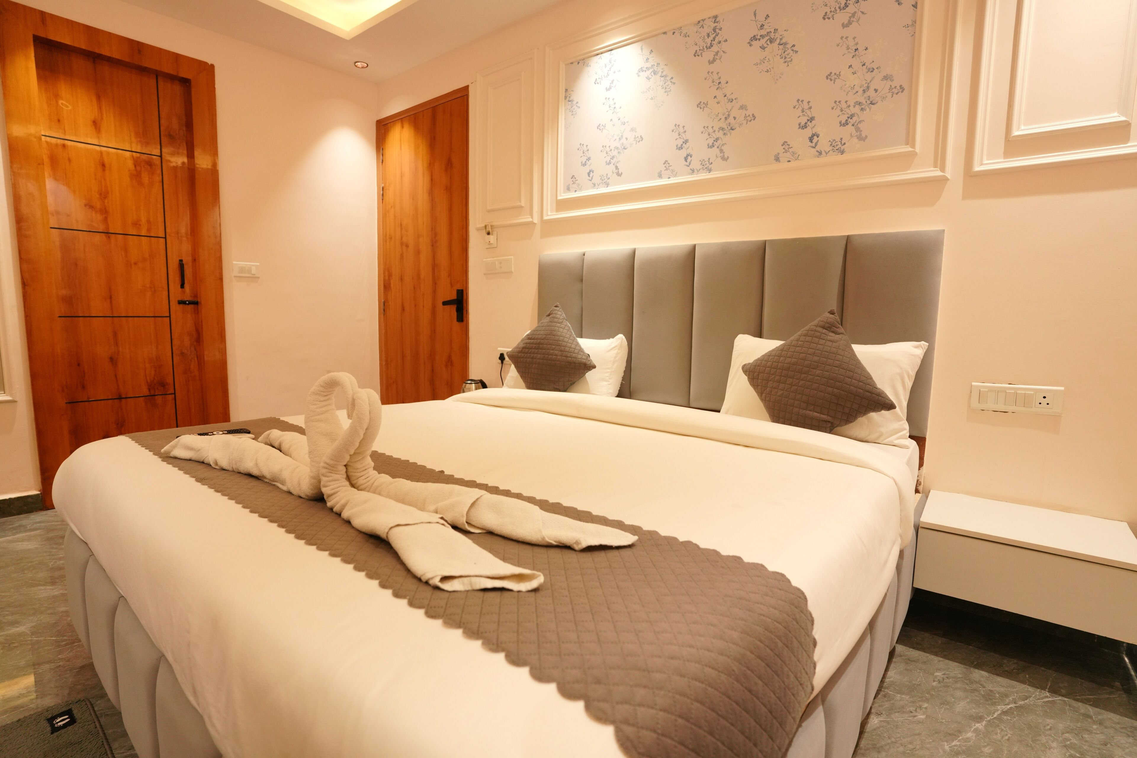 A hotel room with a large bed, a wooden door, a bedside table, and a wall-mounted light fixture.