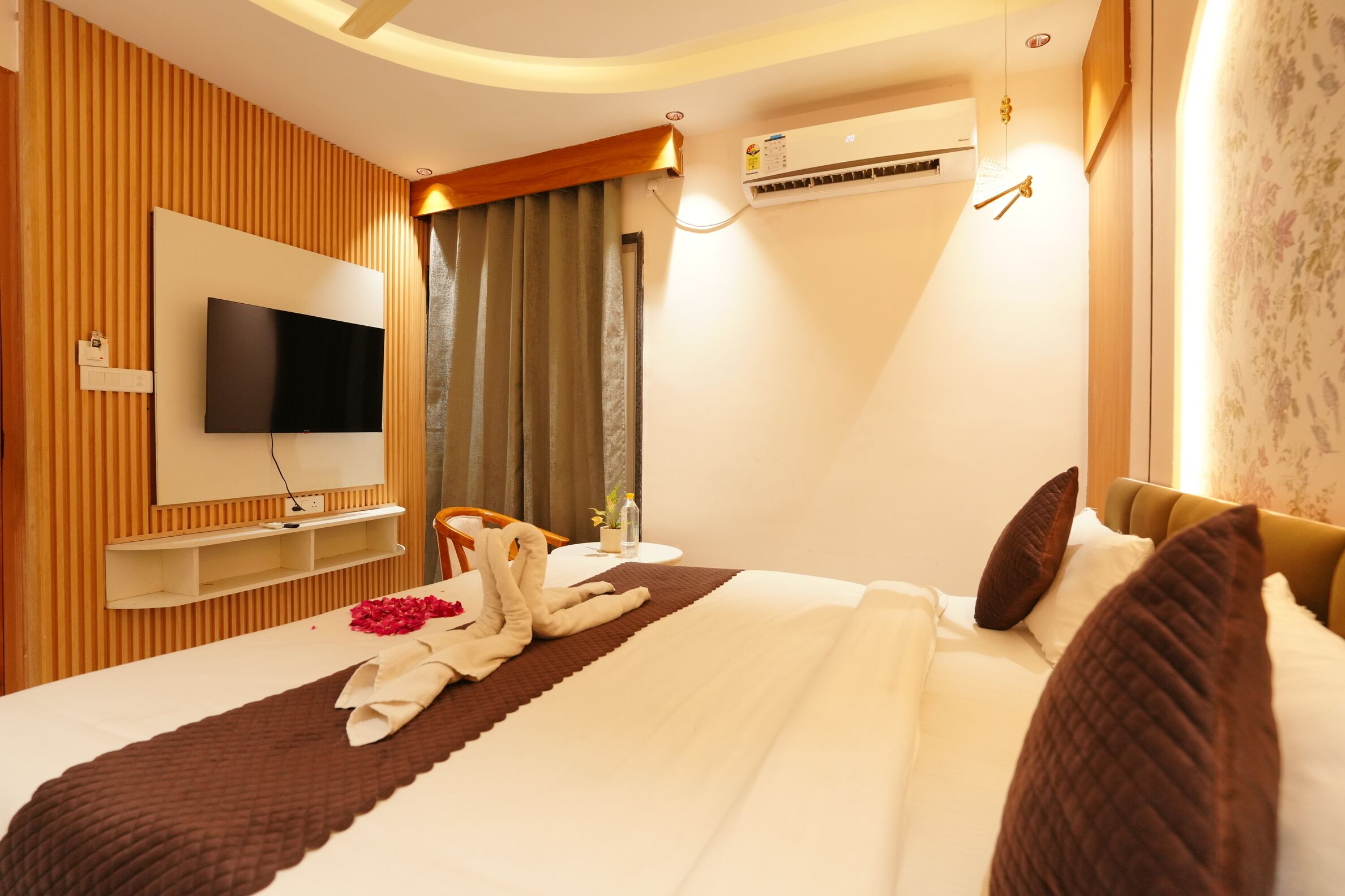 A hotel room with a flat-screen TV mounted on the wall, a bedside table with a bottle and a plant, a bed with a brown blanket and pillows, and an air conditioning unit on the wall.