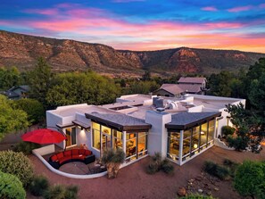 A modern house with a large glass facade, a covered outdoor seating area, and a view of the surrounding landscape. - Hot Tub + Covered Dining + Grill | Red Rock Views | Sage & Sandstone (Sedona)