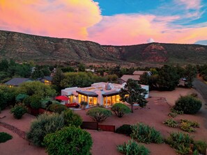 A modern house with a spacious patio, surrounded by lush greenery and a mountainous backdrop. - Hot Tub + Covered Dining + Grill | Red Rock Views | Sage & Sandstone (Sedona)