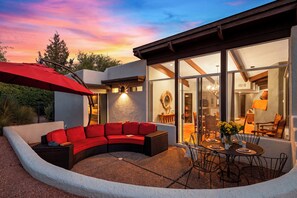 A modern outdoor living space with a curved red sofa, a dining table with chairs, and a large umbrella. - Hot Tub + Covered Dining + Grill | Red Rock Views | Sage & Sandstone (Sedona)