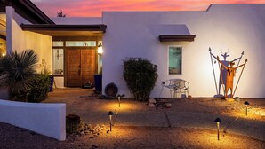 Una casa moderna con facciata bianca, una porta in legno e un piccolo finestrino. Si vedono un cactus e una sedia all'esterno.