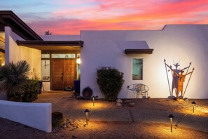 A modern house with a white exterior, a wooden door, and a small window. There is a cactus and a chair outside. - Hot Tub + Covered Dining + Grill | Red Rock Views | Sage & Sandstone (Sedona)