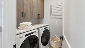 A laundry room with two Whirlpool washers, a wooden cabinet with two doors, a towel rack, and a basket. - Private Pool + Hot Tub + Game Room + Gym | Hill Country Views | River Bend (Austin)