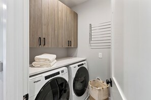 A laundry room with two Whirlpool washers, a wooden cabinet with two doors, a towel rack, and a basket. - Private Pool + Hot Tub + Game Room + Gym | Hill Country Views | River Bend (Austin)