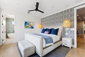 Ein modernes Schlafzimmer mit Bett, Nachttischen, Deckenventilator und einem durch eine offene Tür einsehbaren Badezimmer.