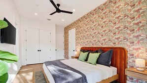 A bedroom with a bed, a ceiling fan, a nightstand, a lamp, and a wall with a floral pattern. - Private Pool + Hot Tub + Game Room + Gym | Hill Country Views | River Bend (Austin)