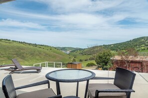 Eine Terrasse mit Glastisch und Korbstühlen, mit Blick auf ein Tal, das ferne Hügel und ein Gewässer zeigt.