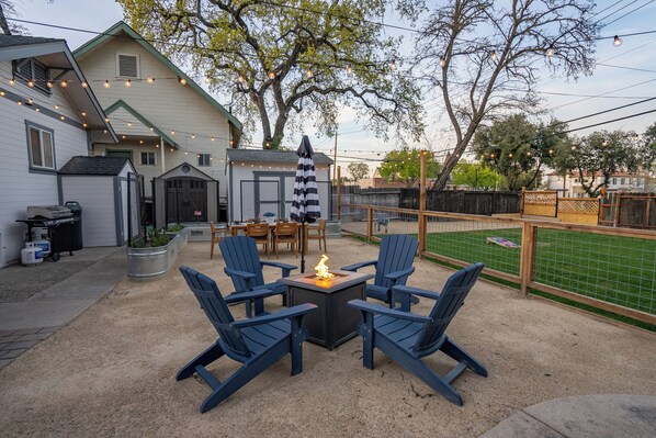Ein Garten mit Feuerstelle, zwei Adirondack-Stühlen, einem Esstisch mit Stühlen und einer überdachten Terrasse.