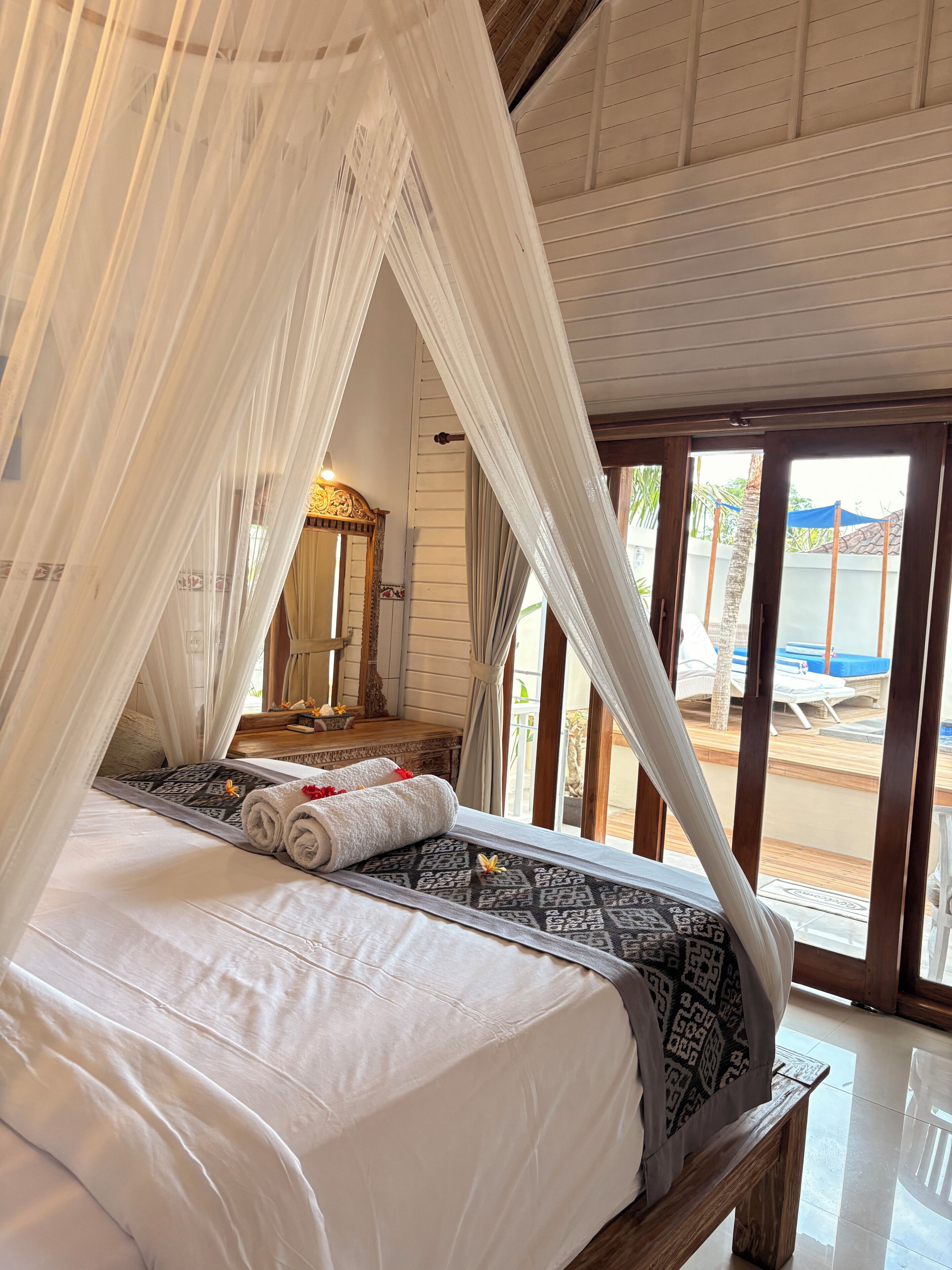 A bed with a canopy, a wooden bench, a mirror, and a view of an outdoor pool area.