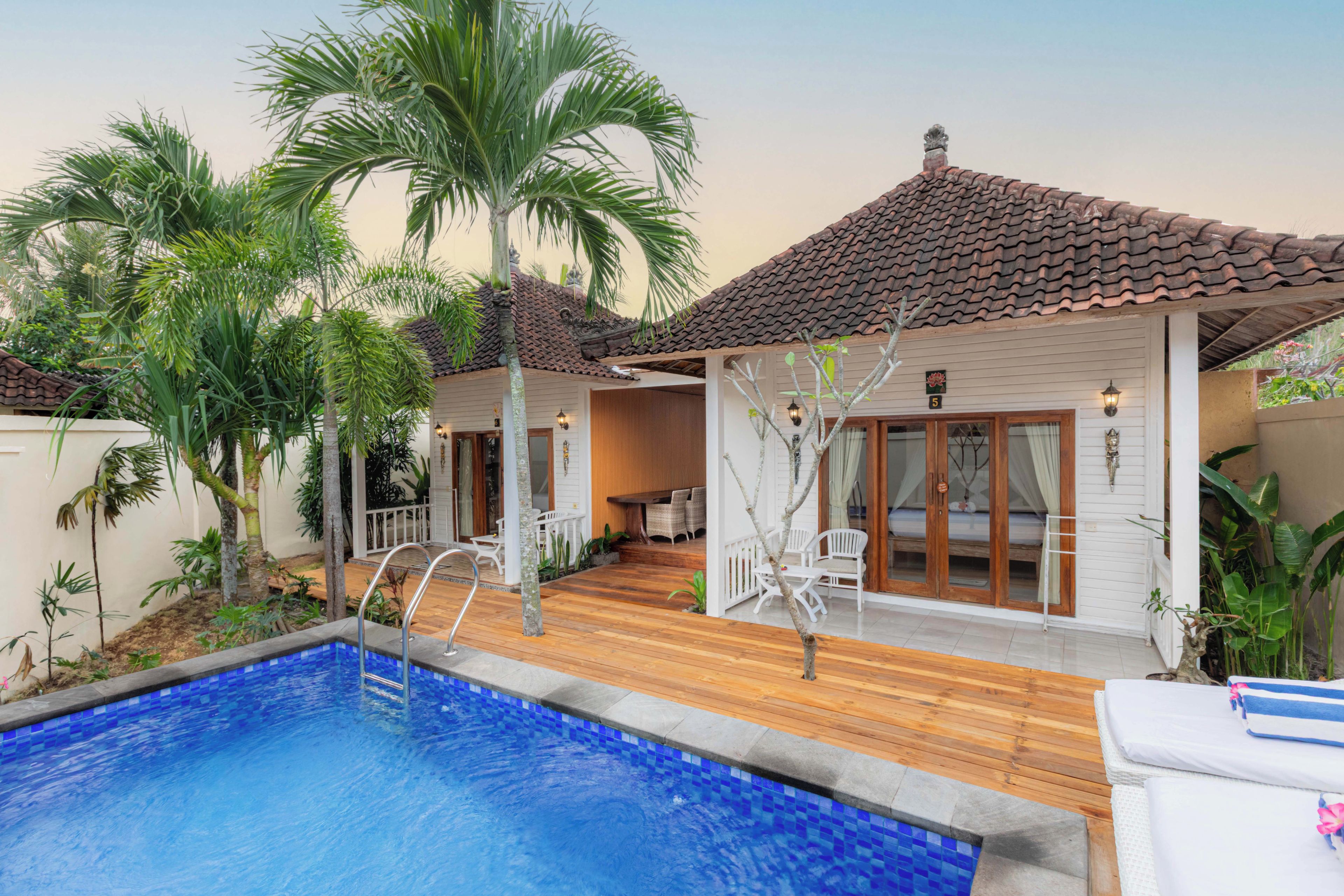 A poolside area with a wooden deck, a swimming pool with blue tiles, and a building with a tiled roof.