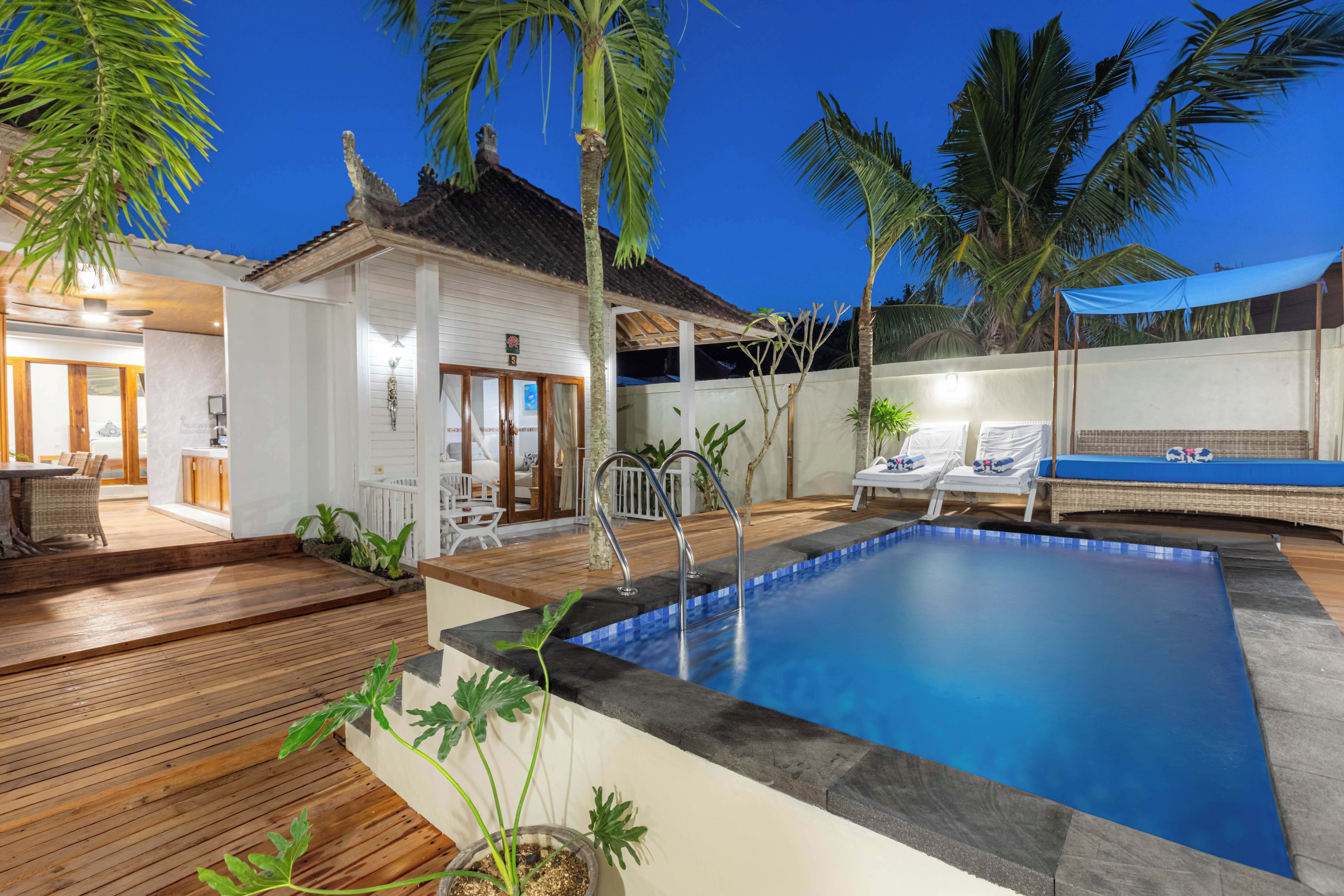 Uma área de piscina com deck de madeira, cadeiras de descanso e um pequeno edifício com telhado ladrilhado.