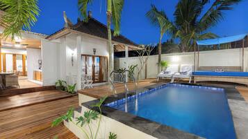 Uma área de piscina com deck de madeira, cadeiras de descanso e um pequeno edifício com telhado ladrilhado.