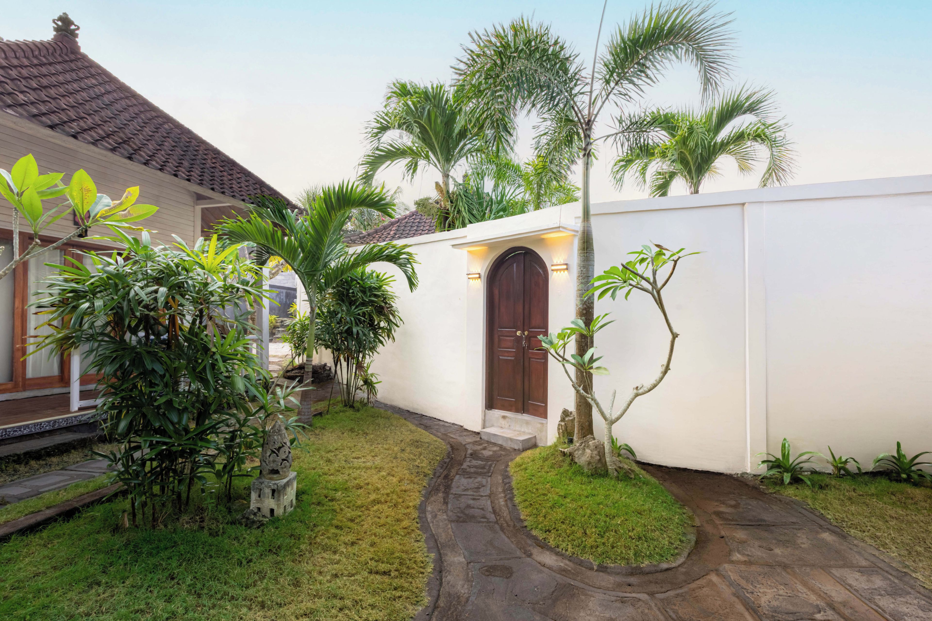 Uma casa tradicional com telhado de azulejos, porta de madeira e um pequeno jardim com palmeiras e um caminho.