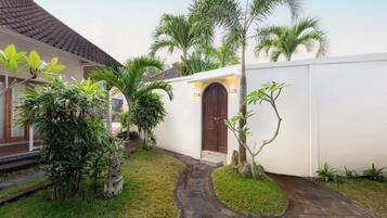 Uma casa tradicional com telhado de azulejos, porta de madeira e um pequeno jardim com palmeiras e um caminho.