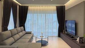 Una sala de estar moderna con un sofá gris, una mesa de centro de vidrio, un televisor de pantalla plana y un ventanal con cortinas translúcidas.