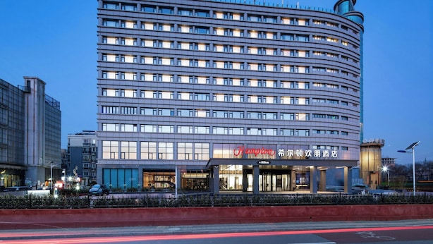 A multi-story hotel with a modern design, featuring a prominent curved corner and a rooftop structure. - Hampton by Hilton Beijing Dahongmen (Beijing)