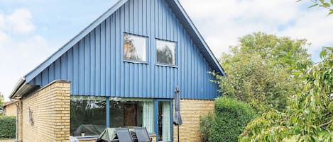 Une maison bleue avec des fondations en briques, un patio couvert avec des meubles d’extérieur et un jardin où poussent différentes espèces de plantes.