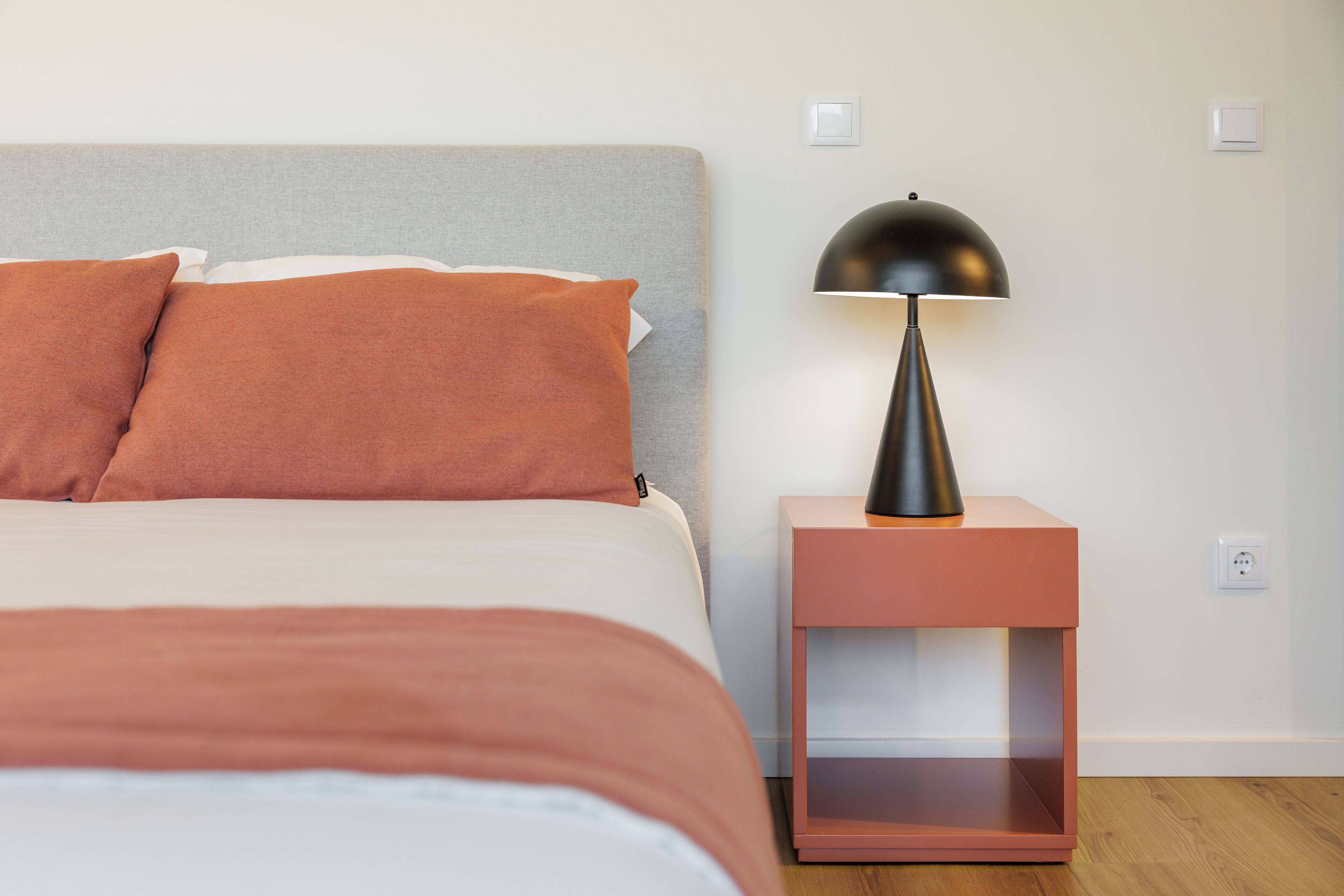 A neatly made bed with a grey headboard and two orange pillows, a black lamp on a pink nightstand, and a white wall with two switches.