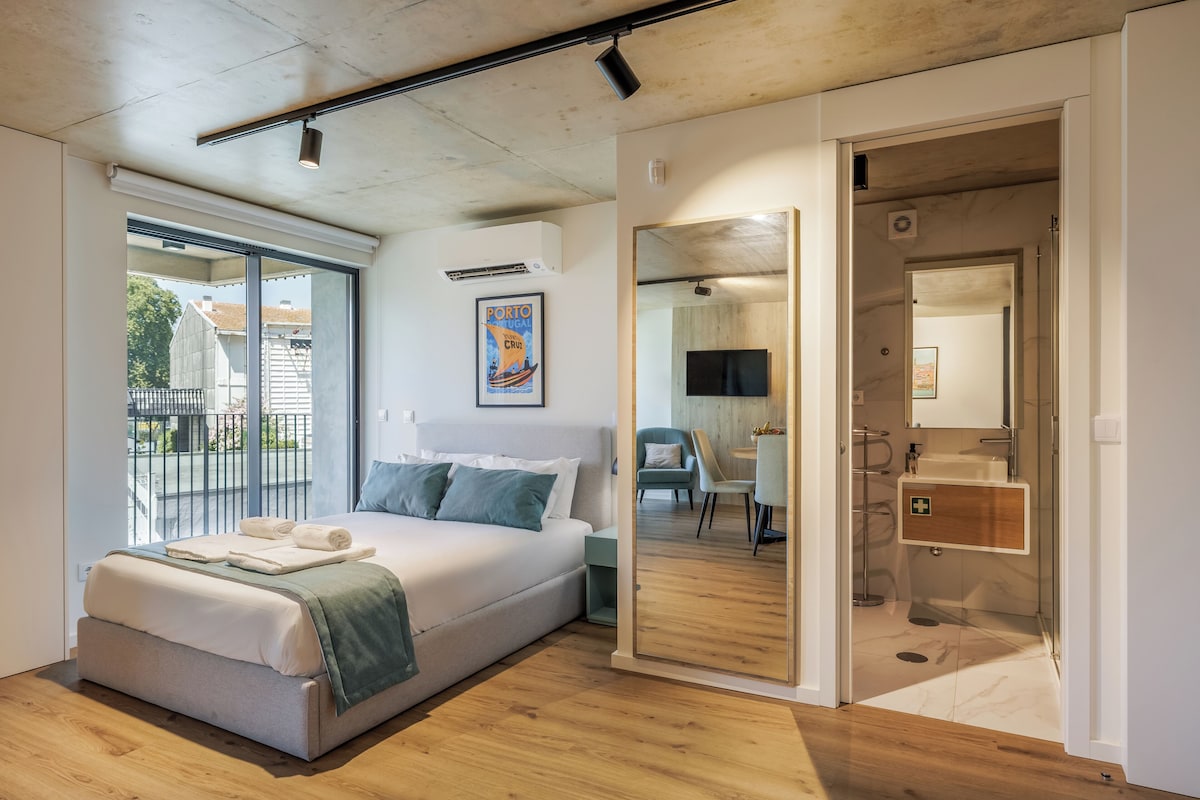 A modern bedroom with a large bed, a view of the outdoors, and a bathroom with a mirror and sink.