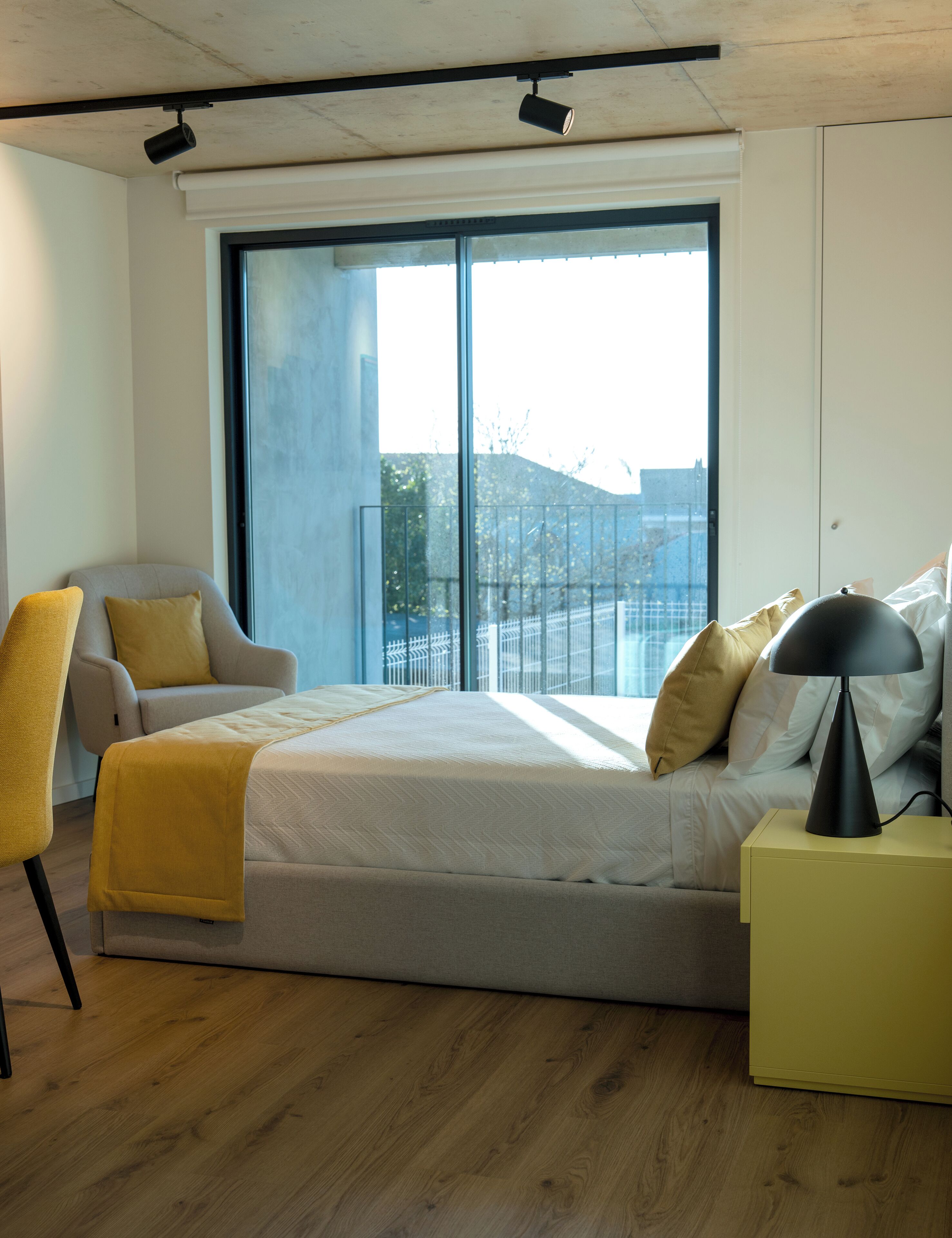 A modern hotel room with a large bed, a chair, a bedside table, and a view of the city through large windows.