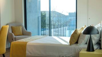 A modern hotel room with a large bed, a chair, a bedside table, and a view of the city through large windows.