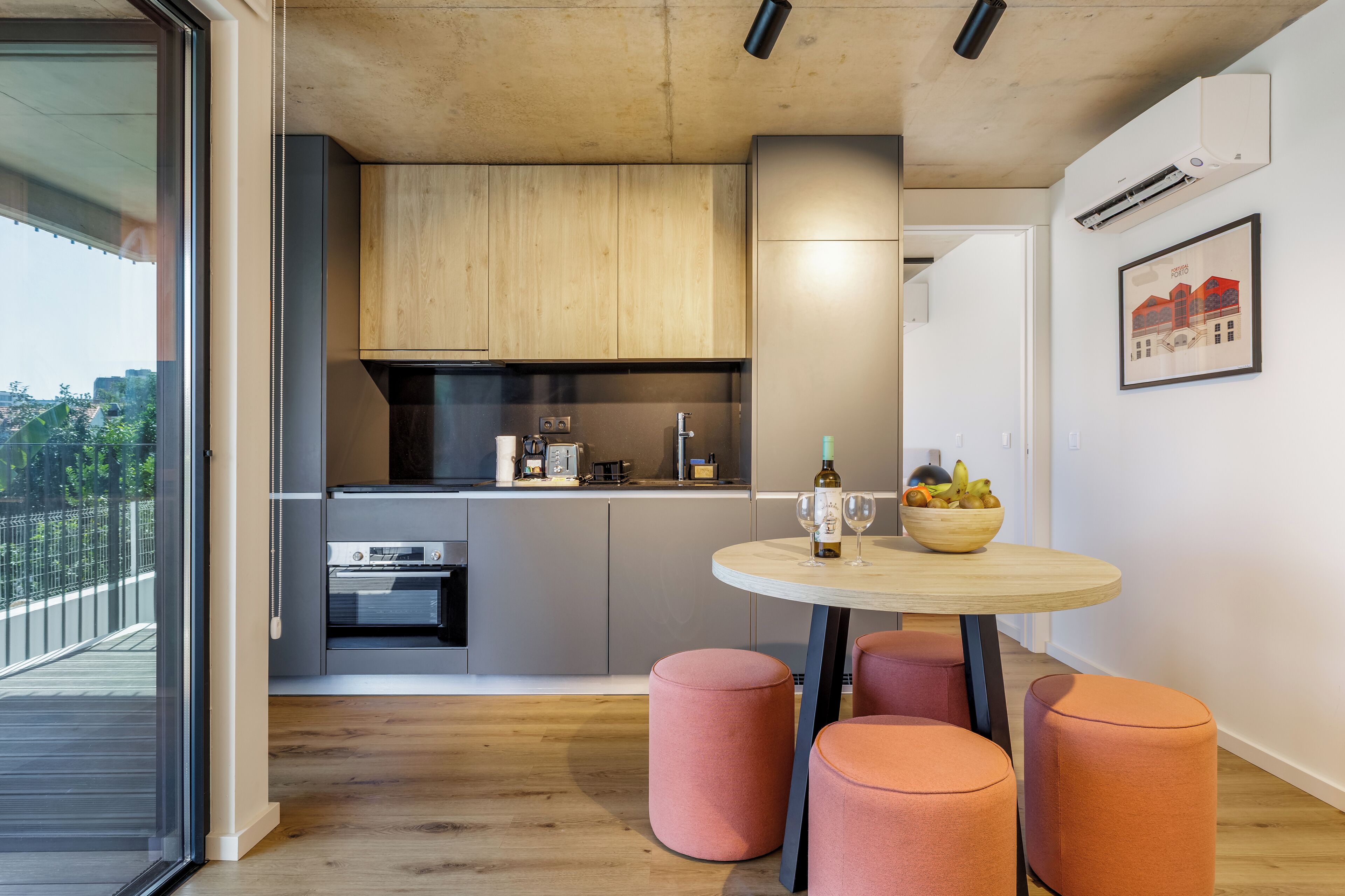 A modern kitchen with wooden cabinets, a built-in oven, and a dining area with a round table and pink ottomans.