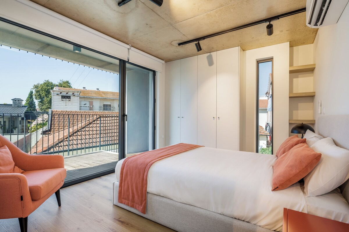 A modern bedroom with a large bed, a chair, and a balcony with a view of buildings.