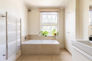 A bathroom with a bathtub, a window with plants, and a towel rack. - Stylish Notting Hill Home with Patio (London)