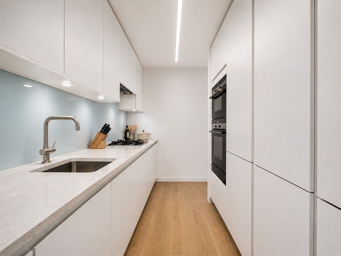 A modern kitchen with white cabinets, a stainless steel sink, a built-in oven, and a wooden floor.