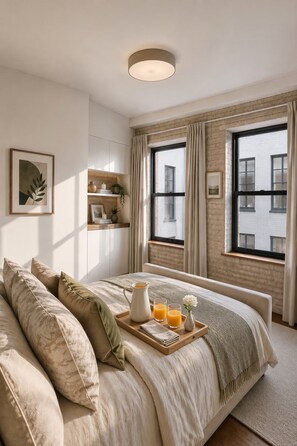 Quarto com uma cama grande, uma mesinha de cabeceira com um bandeja de suco de laranja e vista para um edifício de tijolos pela janela.