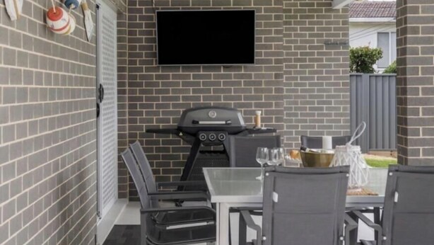 A patio area with a brick wall, a flat-screen TV mounted above a grill, and a dining table with chairs.