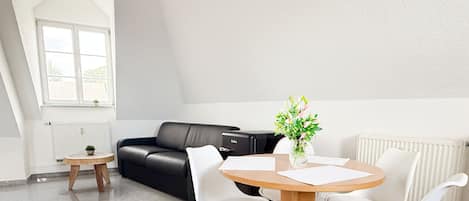 Un salon moderne avec un canapé en cuir noir, des fauteuils blancs, une table basse en bois et un vase rempli de fleurs.
