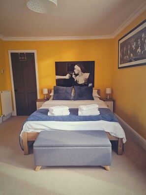 Ein Schlafzimmer mit einem Bett, Kissen, einer Bank, einem Nachttisch, einer Lampe und einem gerahmten Bild an der Wand.