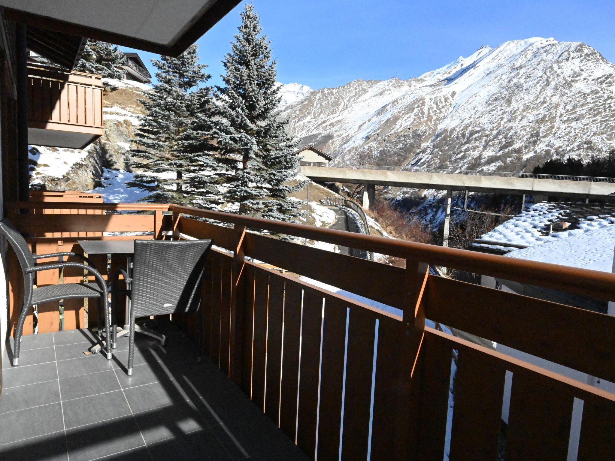 Un balcon offrant une vue sur des montagnes enneigées, avec une balustrade en bois et une table entourée de chaises.