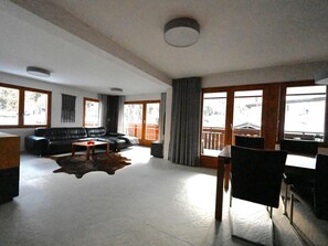 Ein großzügiger Wohnbereich mit einer schwarzen Ledersofa, einem Couchtisch und einem Esstisch mit Stühlen. Große Fenster sorgen für natürliches Licht und einen Blick nach draußen.