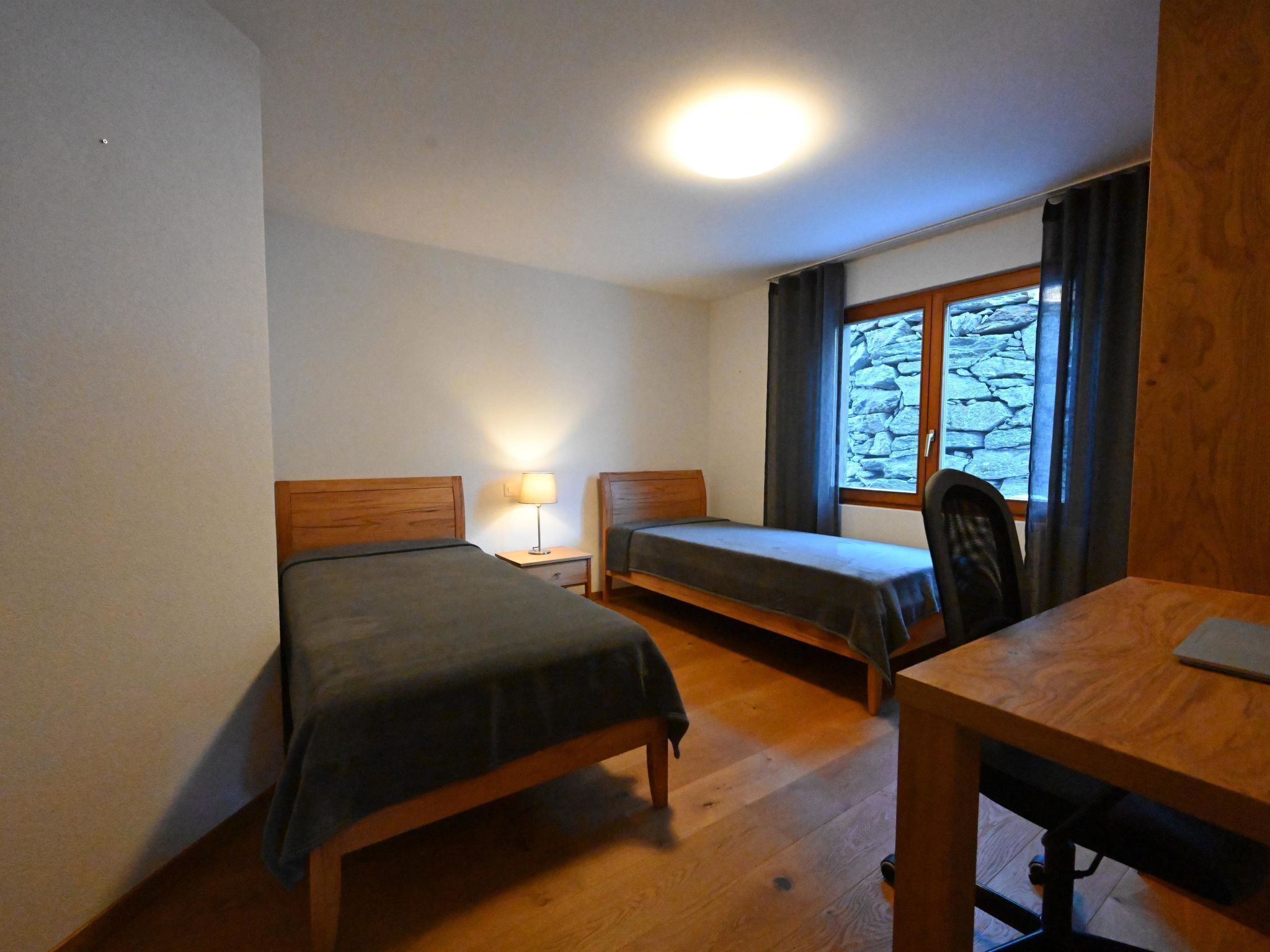 Una habitación con dos camas individuales, un escritorio, una silla, una lámpara y una ventana que da a un muro de piedra.