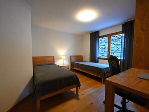 Een kamer met twee eenpersoonsbedden, een bureau, een stoel, een lamp en een raam met uitzicht op een stenen muur.