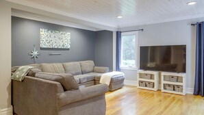En moderne stue med en hjørnesofa, et fladskærms-TV på et hvidt skab og et stort vindue med gardiner.