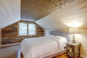Ein Schlafzimmer mit schrägen Wänden, einer Holzwand, einem Fenster, einem Bett und einem Nachttisch mit Lampe.
