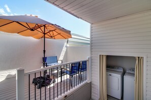 Ein Balkon mit weißer Brüstung, einem blauen Stuhl, einer weißen Waschmaschine und einem großen Sonnenschirm.
